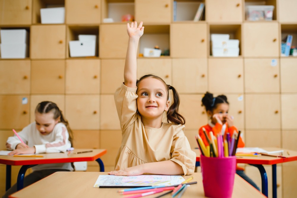 little girl with pigtails raising her hand to ask a question in a classroom